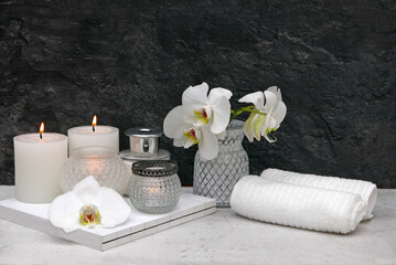 Spa Arrangement mit Körperpflegeprodukten, Badezusatz, Aromaöl und Handtuch vor einer schwarzen Wand mit Platz für Text.