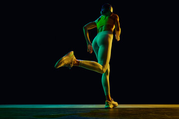 Naklejka premium Wellness. Yung sportive woman in sportswear and headphones in motion, running on black background in neon light. Concept of active and healthy lifestyle, sport, hobby, motivation, endurance