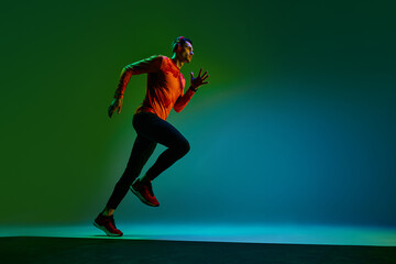 Young athletic man with muscular body, in headphones, training, running on green background in neon...