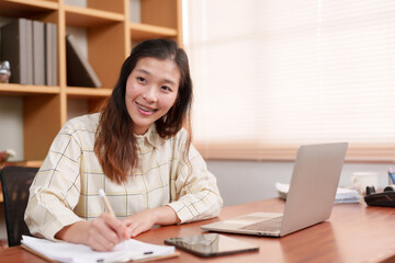 Smiling Asian female sits at a desk, laptop open, pen in hand on notepad, indicating productivity...
