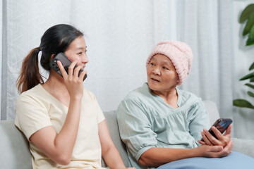 Younger Asian female converses on cellphone, elder in cozy hat handles smartphone, familial bond. Conversation flows as young lady speaks on phone, senior in handmade cap with mobile device - obrazy, fototapety, plakaty