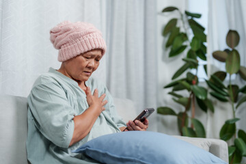 Asian elder with knitted cap displays distress, hand on chest, gripping phone,  Woman in cancer...