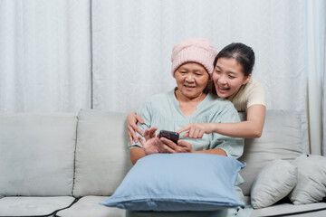 Cheerful Asian woman in cancer headwear explores smartphone, younger companion leaning in, Joyous...