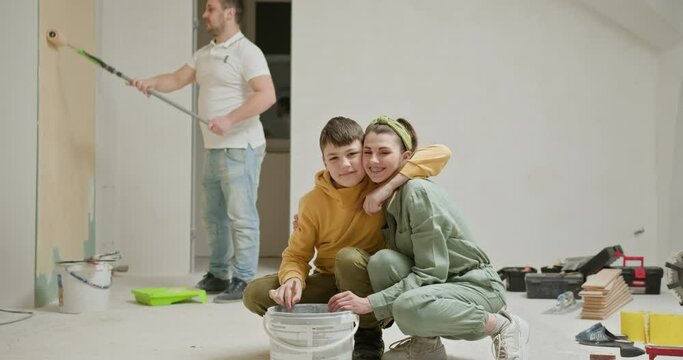 A heartwarming scene of a family engaged in home renovation. Mom and son smiling near a paint bucket while dad paints the wall behind them. Joy of family teamwork and the steps of DIY home improvement