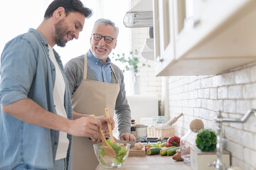 Cheerful adult son helping his senior old father in cooking, preparing family meal, dinner, lunch. Parents visiting. Happy father`s day! I love you, dad! Men cooking together - Powered by Adobe