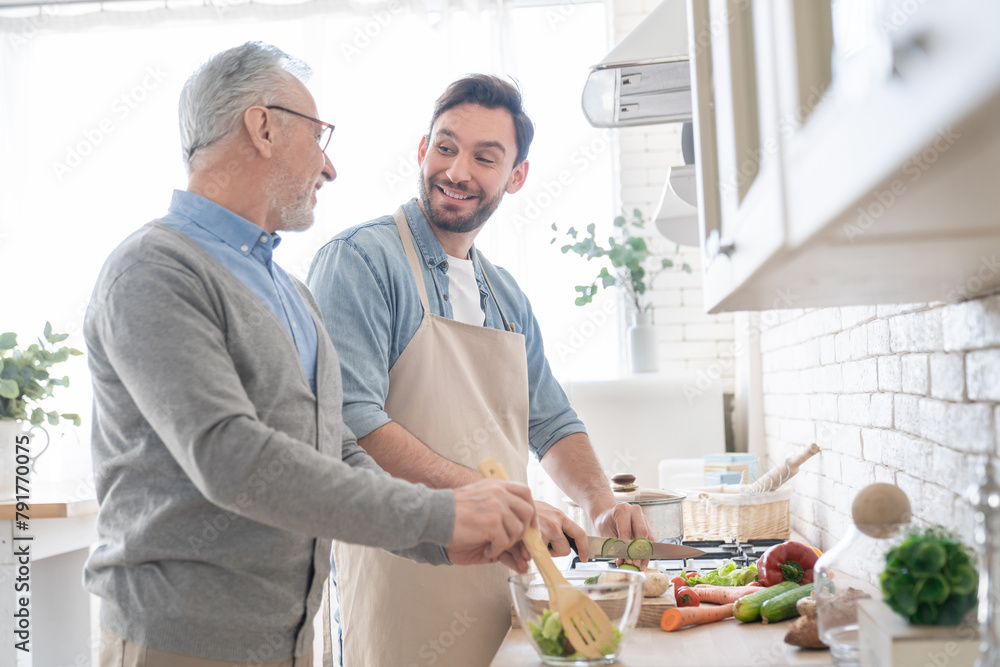 Wall mural happy young caucasian son talking with his old elderly senior father while cooking dinner, lunch in 