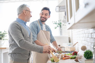 Happy young caucasian son talking with his old elderly senior father while cooking dinner, lunch in...