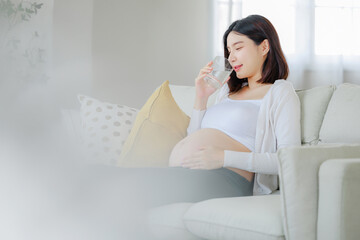 Pregnant woman drinking water sitting on sofa. Young expectant lady have rest at home with glass,...