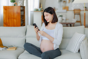 Young Asian happy pregnant woman is sitting and using mobile phone on sofa and touching her belly....