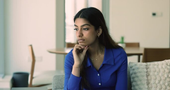 Thoughtful young attractive Indian woman looking away in deep thoughts, thinking on problems, bad troubles, making serious decision, leaning chin on hand, sitting alone at home couch