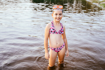 Happy preteen girl in swimsuit and mask in lake water standing knee-deep in water. Summertime...