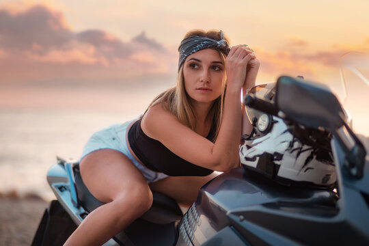 Portrait of pretty young woman poses sitting on motorcycle. Golden sunset and motorbike on the background. Concept of World Motorcyclist Day and moto travel
