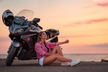 Smiling young woman sitting and posing near motorcycle. Golden sunset and motorbike on the...