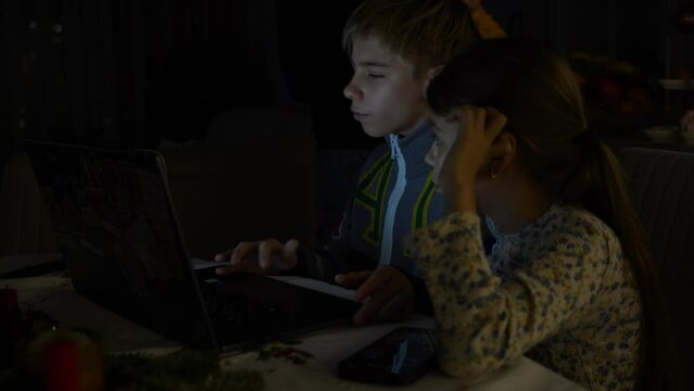 Two children are intently trying to complete school assignments on a laptop, but they are constantly distracted by the TV in a dimly lit room