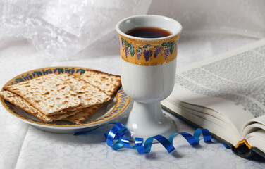 Passover with a goblet of wine, unleavened bread on a plate, and an open book. The goblet is ornate and the bread is matzah. A blue ribbon is near the book on a textured cloth in the background.
