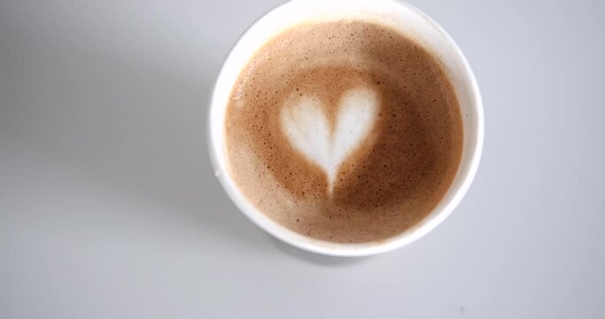 On the table there is a disposable recyclable eco cup in which hot cappuccino coffee with foam and a heart pattern is poured. Coffee with alternative oat milk. A morning cup of invigorating drink.