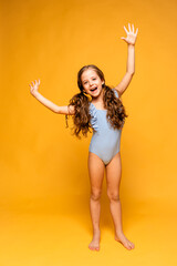 A beautiful cute little girl in a blue swimsuit laughs cheerfully, raise her hands up on yellow...