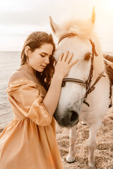 A woman in a dress stands next to a white horse on a beach, with the blue sky and sea in the...