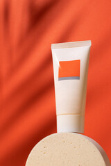 White plastic tube with face, hand and body cream on podium on orange background with palm leaves shadow. Sun protection lotion, sunscreen. Summer skin care concept with spf. Mockup
