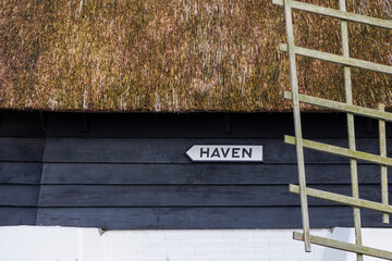 A signpost sign with the Dutch text Harbour on a dark painted wooden wall under a thatched roof of a polder windmill for draining the ditches in a residential area