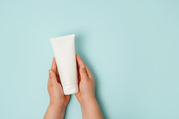 Child hands holding white cream tube on light blue background. Care about clean and soft body skin....