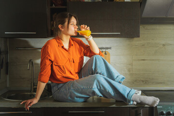 Beautiful young woman sitting on a tabletop with a glass of orange juice. - 791838868