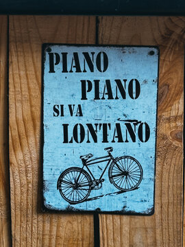 A sign with a bicycle on it that says "Piano Piano Siva Lontano". The sign is blue and white