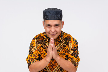 An honest and friendly Indonesian man wearing a patterned batik shirt and a songkok is seen with hands together offering a prayer or greeting. - obrazy, fototapety, plakaty