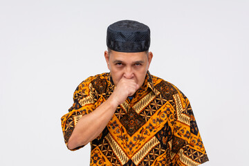 Fototapeta premium An Indonesian man in traditional batik shirt and kopiah hat looking angry while coughing, isolated on a white background.