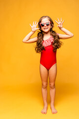 A beautiful cute little girl in a red swimsuit laughs cheerfully and showing her palms hands on yellow background. Mock up. Summer vacation concept. Advertising of children's products and sale