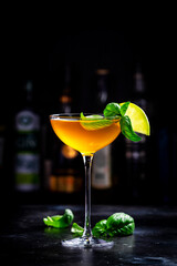 Spring orange cocktail drink with scotch whiskey, apple liqueur, ginger, lemon juice, green basil and ice, dark bar counter background