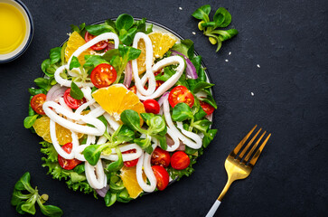 Fresh salad with squid, oranges, cherry tomatoes, lamb lettuce and olive oil with lemon juice dressing, black table background, top view - 791861253