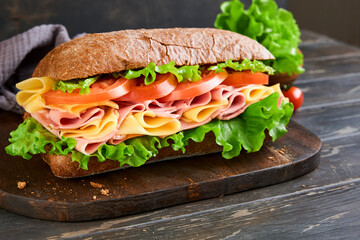 Sandwich. One fresh big submarine sandwich with ham, cheese, lettuce, tomatoes and microgreens on old wooden dark background. Healthy breakfast theme concept, school lunch, breakfast or snack.