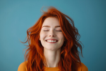 Fototapeta premium Vibrant image capturing a young ginger-haired woman blissfully smiling with her eyes closed, set against a soothing blue backdrop.
