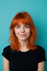 Fototapeta premium A young woman with striking red hair and a subtle smile stands against a vibrant teal background, radiating understated elegance in a classic black top.