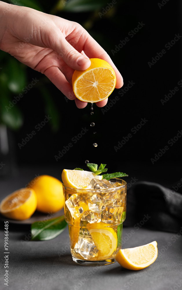 Canvas Prints A hand squeezes juice from a lemon into a glass with lemonade with ice and mint on a dark background with citrus fruits and a branch.