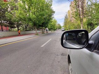 It shows a street scene seen from the perspective of a car's side mirror. The mirror reflects the...