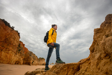 Woman hiker with backpack walking up rocks by a beach, wearing a yellow coat.