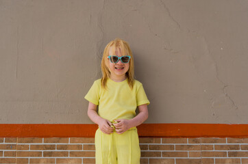 niña rubia usando ropa colorida mientras mira al frente usando lentes junto a una pared gris 