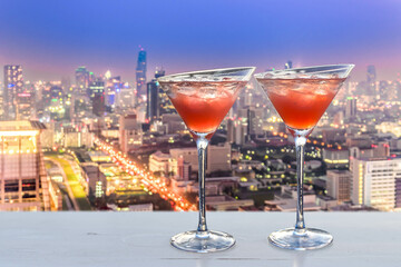 two red cocktails with ice cubes, blurred lights of Bangkok in the background