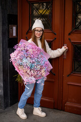 Girl with eyes closed in glasses stands near a wooden door with a bouquet
