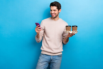 Portrait of positive nice handsome man look at smartphone holding two cups of coffee read notification isolated on blue color background