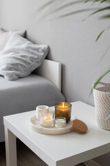 Home decor, stylish interior. Burning candles on marble tray standing on white badside table in bedroom.  Bed with grey blanket and pillows, wicker basket.