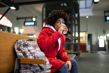 Young curly-haired guy having coffee and waiting for the train at the railway station