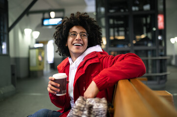 Young curly-haired guy having coffee and waiting for the train at the railway station