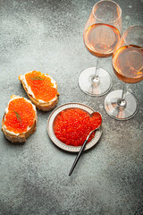 Small metal plate with red salmon caviar, two caviar toasts canape, two glasses of champagne top view on grey concrete background, festive luxury delicacy and appetizer.