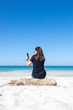 A serene image capturing a lone woman sitting on a log at the beach, gazing out towards the endless blue ocean under a clear sky
