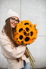Cheerfully girl in knitted hat and sweater with a sunflowers near a wall