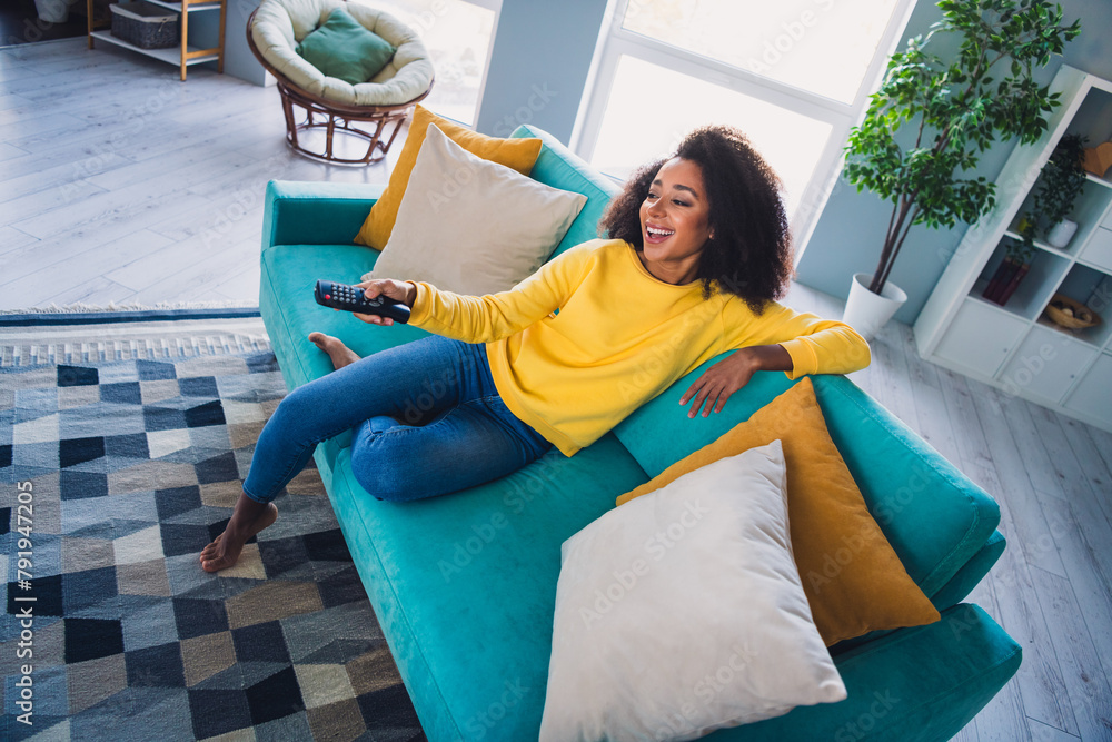 Wall mural Photo of adorable lovely cute girl sitting on sofa turning on tv watching movie in cozy modern apartment