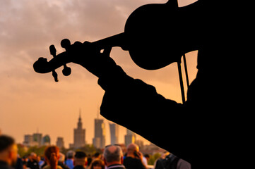Sylwetka, grający na skrzypcach muzyk, zbierający pieniądze na tle Warszawskiego centrum o zachodzie słońca. Zdjęcie z mostu na centrum Warszawy